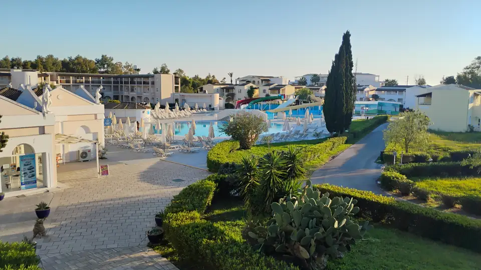 Poolanlage mit Liegestühlen, Wasserrutschen und niedrigen Hotelgebäuden hinter einem begrünten Weg