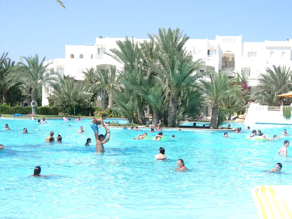 Großer Außenpool mit Badegästen, einer Palmeninsel und weißen Hotelgebäuden