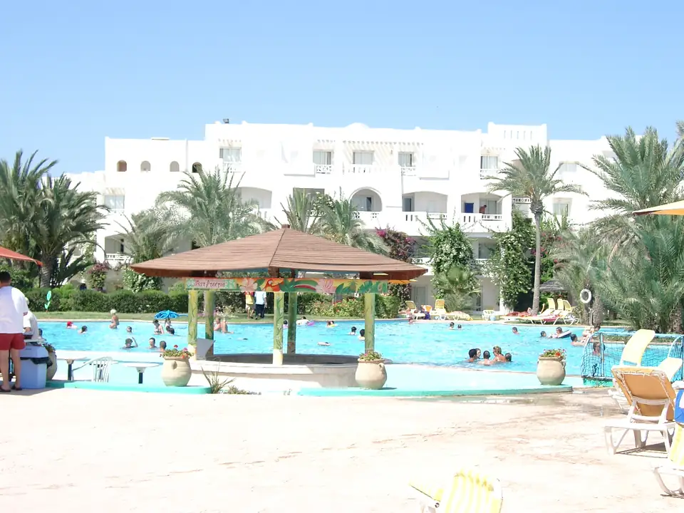 Pool mit einem als Bar Freesia beschrifteten Pavillon vor weißen Hotelgebäuden und Palmen