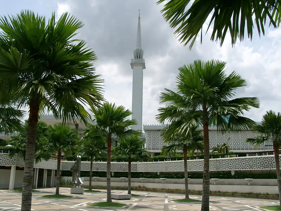 Schlankes weißes Minarett der Nationalmoschee zwischen Palmen und geometrischen Mauerelementen
