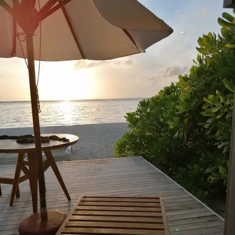 Blick von einer Holzterrasse mit Liege und Sonnenschirm auf den Strand und die Morgensonne über dem Meer