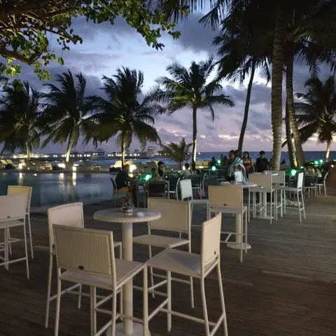 Sitzterrasse mit hellen Stühlen, Palmen und beleuchtetem Pool bei Sonnenuntergang