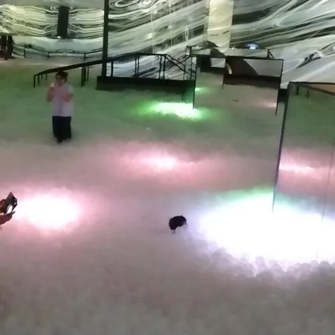 Besucher im weißen Bällebad, umgeben von Spiegelwänden und rosa, grünen und weißen Lichtflecken