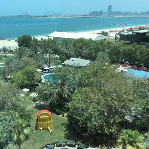 Blick von oben auf den Palmengarten und Pool des Sheraton Jumeirah Beach Resort mit Rasenflächen, Liegen und Strand vor dem türkisfarbenen Meer