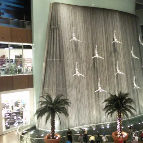 Hoher Wasserfall in der Dubai Mall mit silbernen Figuren in Kopfsprungpose und Palmen davor