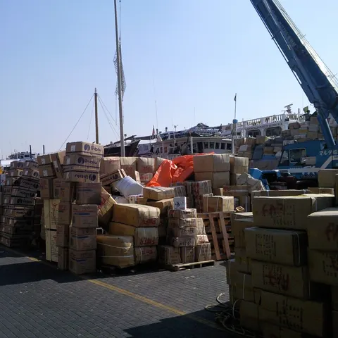 Hohe Stapel von Frachtkartons und Warenballen am Dubai Creek mit Dhaus und Kränen dahinter