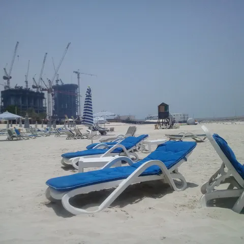 Weiße Strandliegen mit blauen Auflagen und gestreiften Sonnenschirmen im Sand, dahinter ein Rettungsschwimmerturm und Baukräne