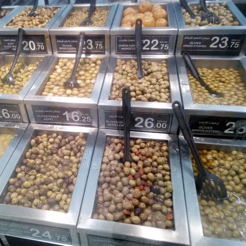 Verschiedene Olivensorten und eingelegte Zitronen in offenen Edelstahlbehältern mit Schöpfkellen und Preisschildern