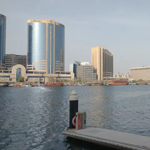 Blick über den Dubai Creek auf Hochhäuser und hölzerne Dhaus am gegenüberliegenden Ufer, mit schwimmenden Anlegestegen im Vordergrund