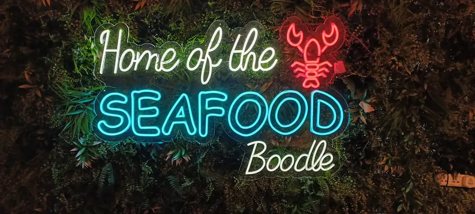 Leuchtschrift „Home of the Seafood Boodle“ mit rotem Hummermotiv vor einer begrünten Wand im La Mesa Restaurant