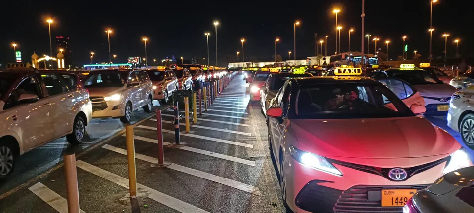 Lange Reihen wartender Taxis vor Global Village bei Nacht, getrennt durch markierte Fahrspuren und Poller