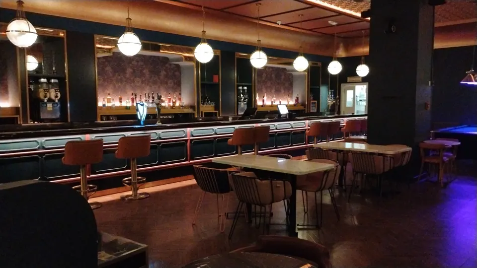 Lange Bar bei Swingers Dubai mit gepolsterten Barhockern, runden Hängeleuchten und Tischen im Vordergrund