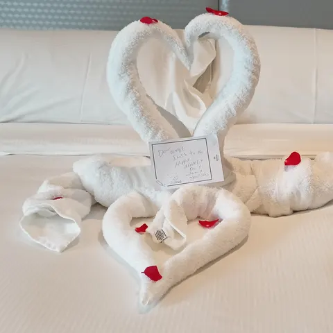 Zwei Handtuchschwäne bilden ein Herz auf dem Bett, davor liegen weitere Handtuchfiguren mit roten Blüten