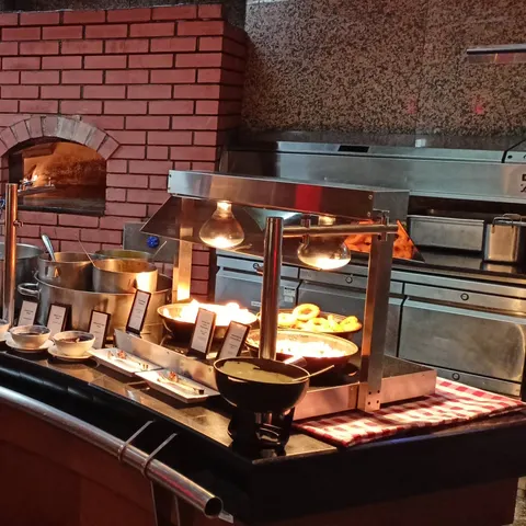 Warme Buffetstation mit beschrifteten Schalen, Wärmelampen und einem gemauerten Ofen im Hintergrund