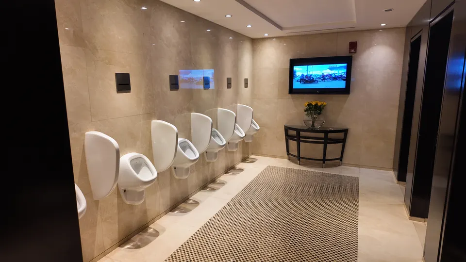 Leere Herrentoilette im Grosvenor House mit einer Reihe weißer Pissoirs und einem Fernseher an der Seitenwand