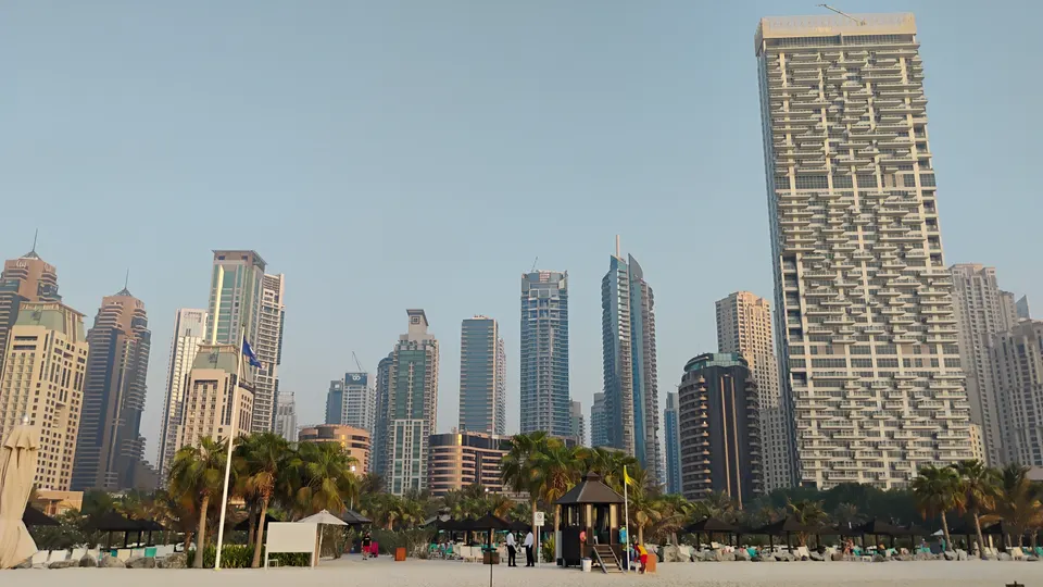 Blick vom Sandstrand auf Holzpavillons und Palmen des Le Royal Méridien mit den Hochhäusern der Dubai Marina dahinter
