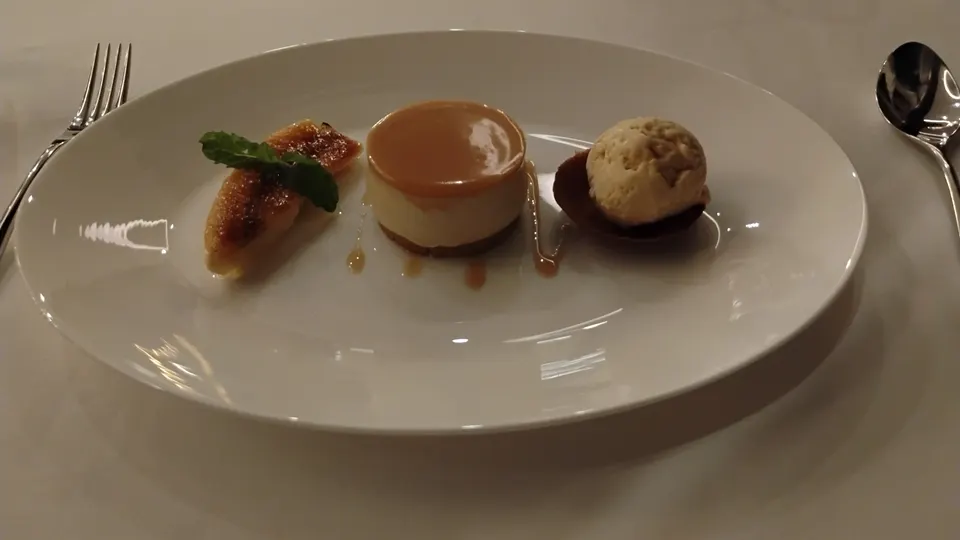 Dessert auf einem großen weißen Teller mit einem runden Cremetörtchen, karamellisierten Bananenstücken, Minze und einer Kugel Eis