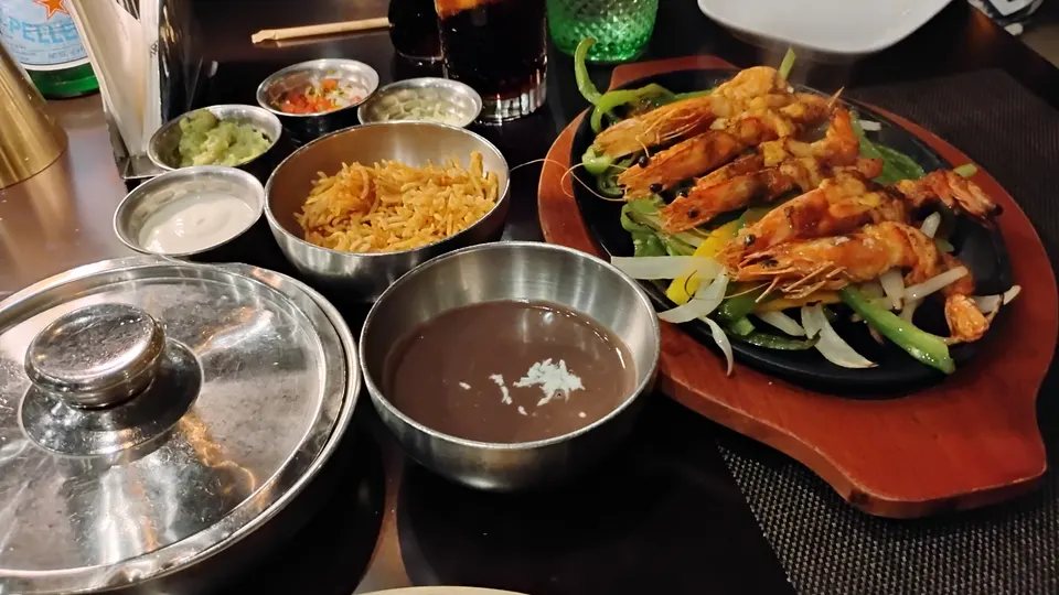 Garnelen auf Paprika und Zwiebeln in einer heißen Platte, daneben Reis, Bohnenmus und kleine Schalen mit Dips