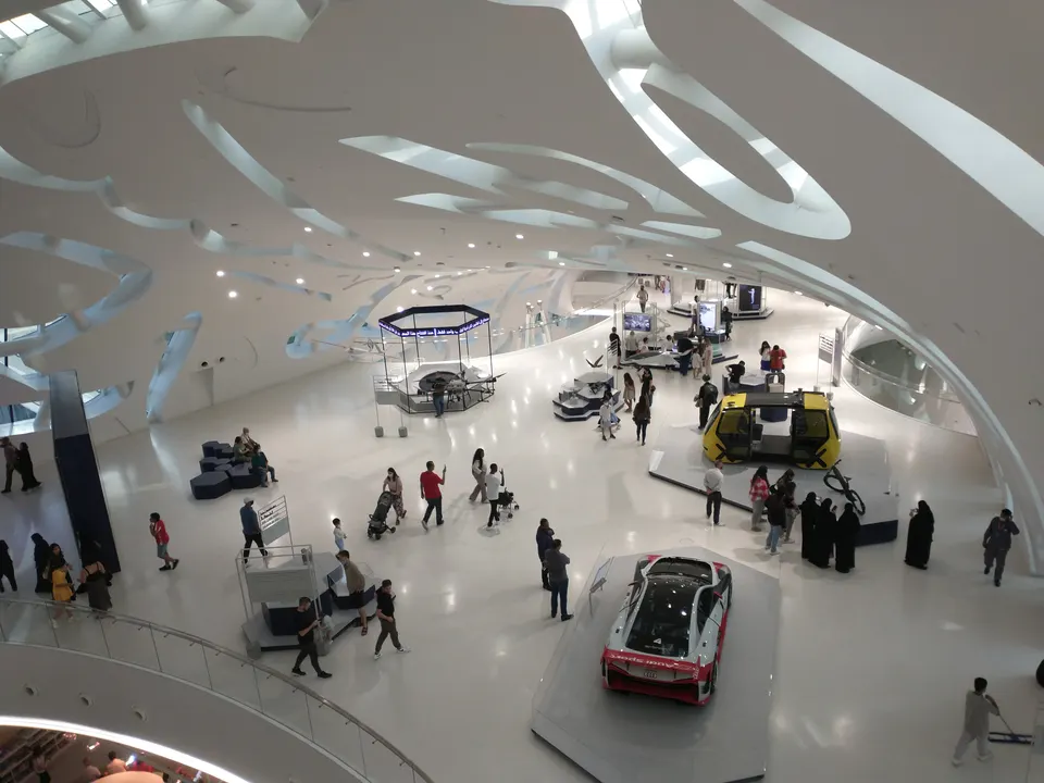 Blick von oben auf einen weißen Ausstellungsraum mit Besuchern, Fahrzeugen und einzelnen Präsentationsflächen