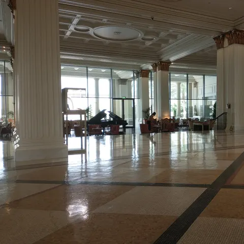 Weitläufige Lobby mit hohen weißen Säulen, glänzendem Mosaikboden und Blick auf einen Flügel vor den großen Fenstern