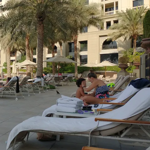 Liegestühle und Sonnenschirme auf der Poolterrasse vor der Hotelfassade und hohen Palmen