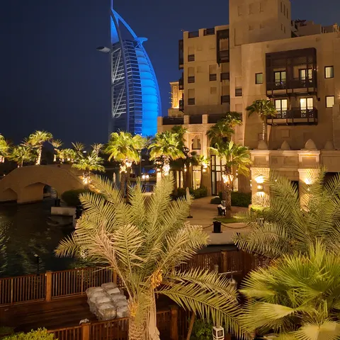 Beleuchtete Gebäude mit Windturm und Palmen im Madinat-Jumeirah-Resort, dahinter der blau beleuchtete Burj Al Arab
