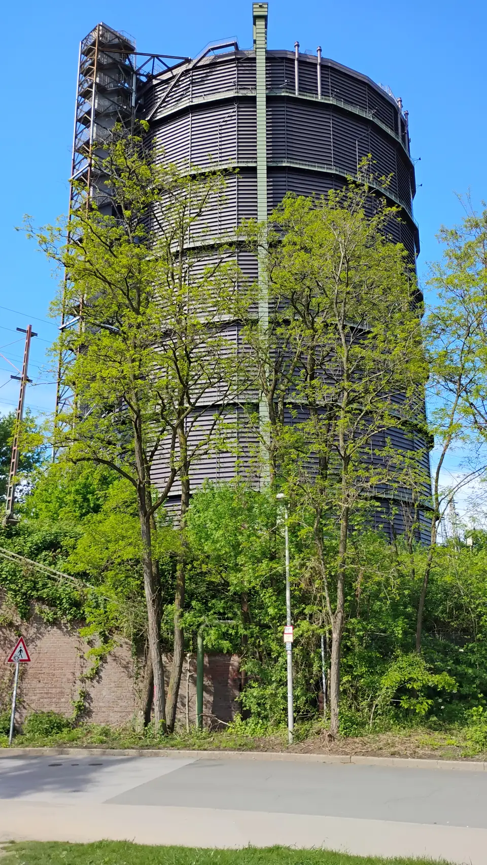 Hoher zylindrischer Gasometer mit außenliegender Treppe hinter Bäumen