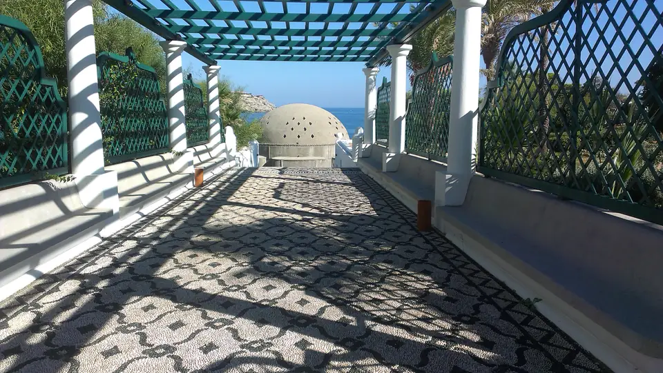Pergolagang mit grünen Gittern, weißen Säulen und Kieselmosaik vor Meer und Rotunde