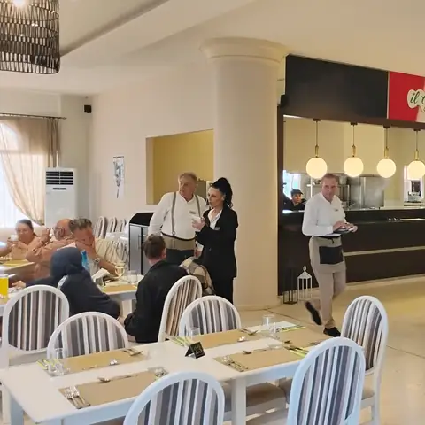 Heller Speisesaal mit weißen Stühlen und einem offenen Küchenbereich unter einem grünen Schild mit der Aufschrift Il Gusto