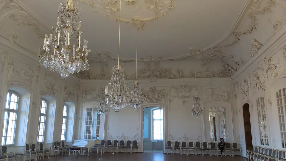 Weißer Ballsaal mit Stuckreliefs, hohen Fenstern und Kristalllüstern