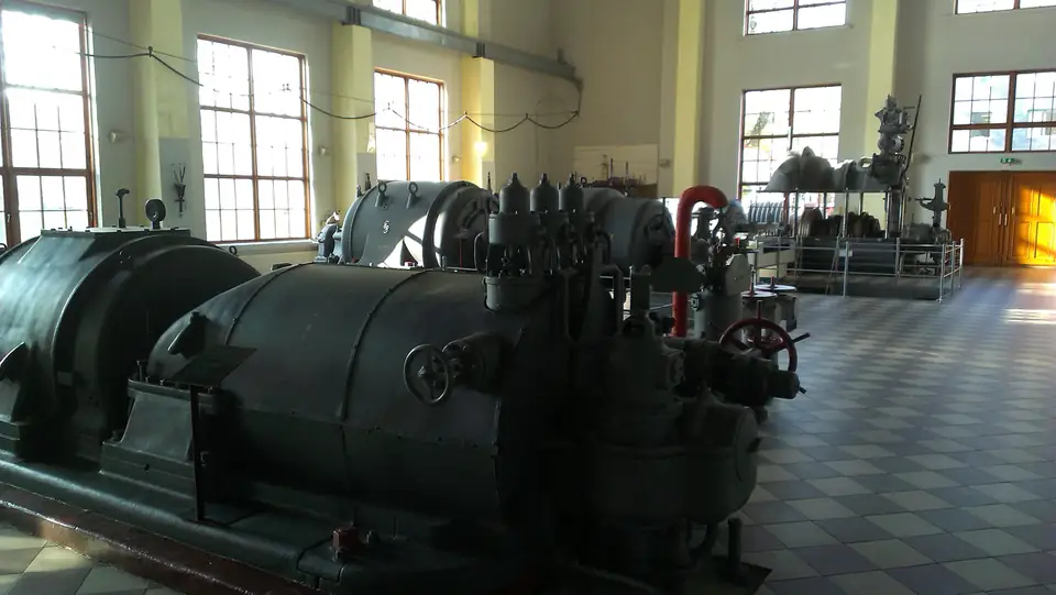 Historische Maschinen mit großen Metallgehäusen und Rohrleitungen in einer hellen Kraftwerkshalle