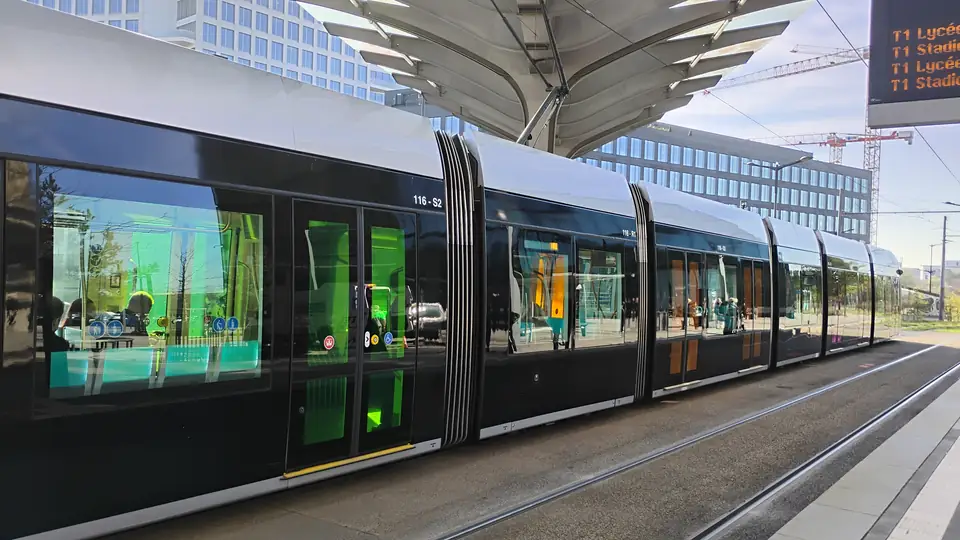 Lange, moderne Straßenbahn an einer überdachten Haltestelle mit Anzeigetafel für die Linie T1