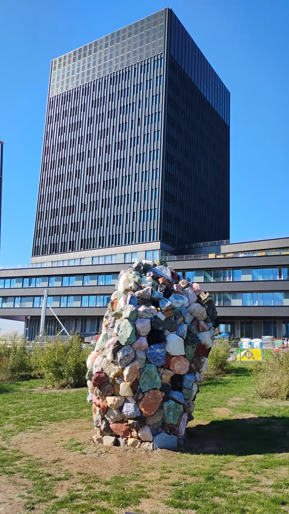 Skulptur aus aufgetürmten bunten Steinen auf einer Wiese vor einem dunklen Hochhaus