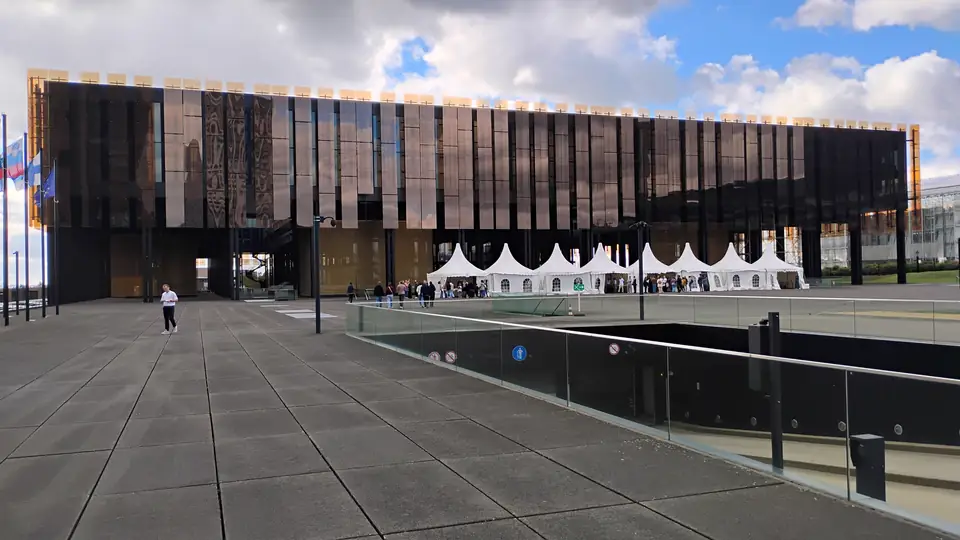 Langgestrecktes Gebäude mit goldbraunen Fassadenlamellen, davor ein weiter Vorplatz mit weißen Zelten