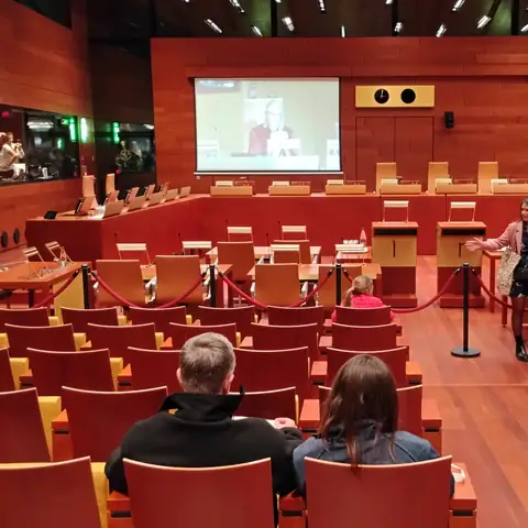 Sitzungssaal mit rot gepolsterten Stuhlreihen, Holzvertäfelung und großer Leinwand
