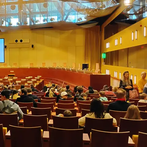 Großer Saal mit voll besetzten Stuhlreihen unter einer goldenen Deckenkonstruktion