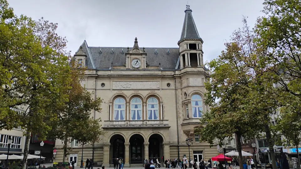 Historisches Gebäude mit Uhrengiebel, Rundbogenfenstern und Eckturm zwischen herbstlichen Bäumen, davor viele Menschen