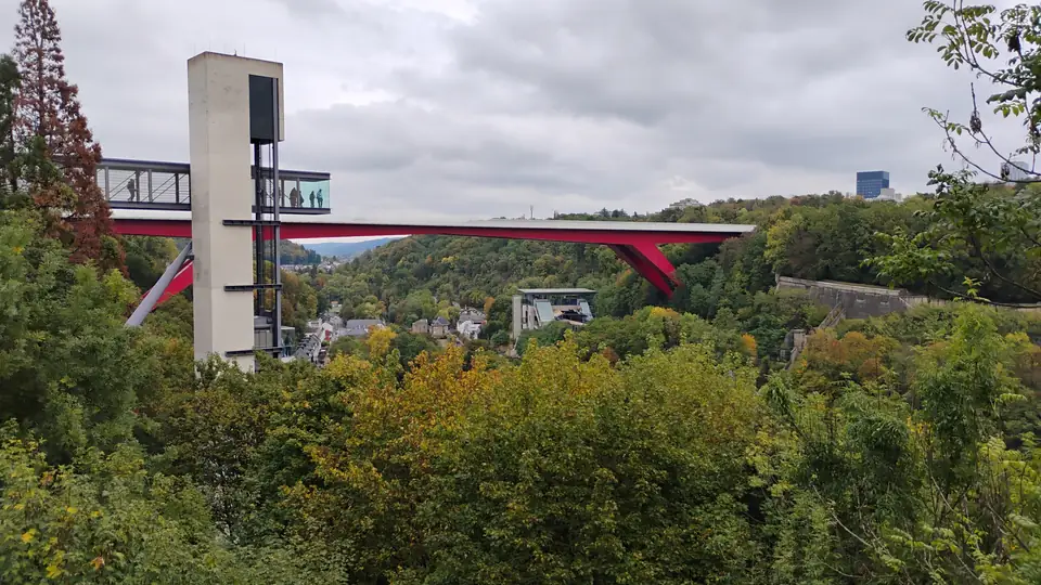 Schlanker Betonturm mit verglaster Aussichtsplattform vor einer roten Stahlbrücke über ein herbstlich bewaldetes Tal
