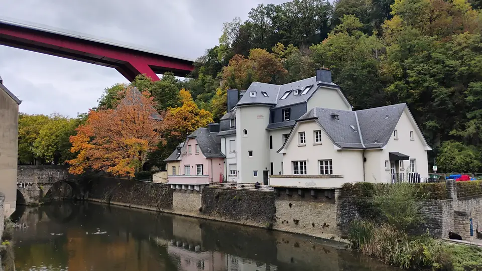 Rote Stahlbrücke hoch über weißen Häusern, herbstlich gefärbten Bäumen und einem ruhigen Fluss