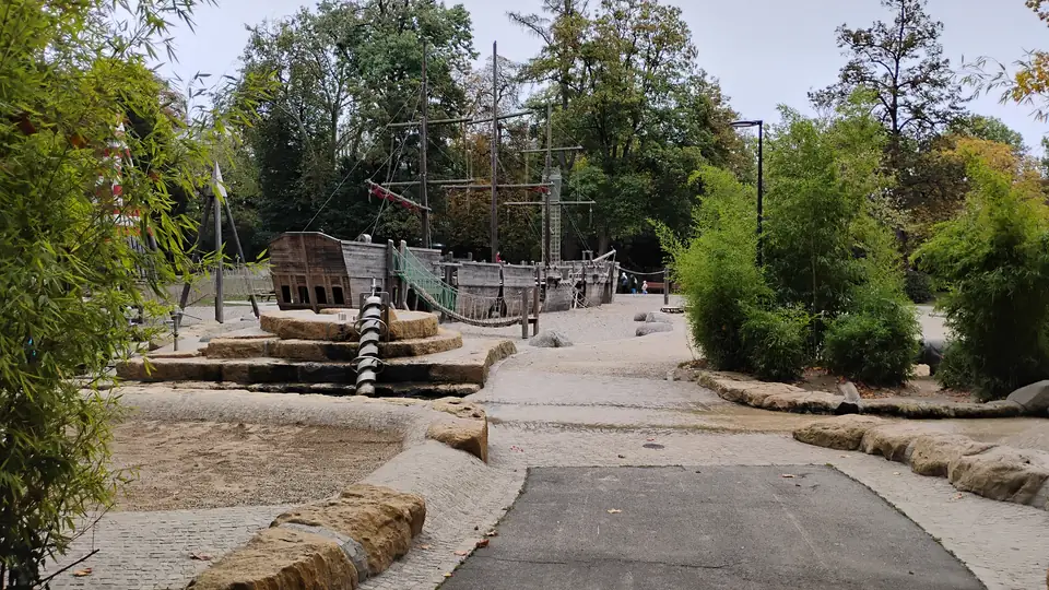 Spielplatz mit hölzernem Schiff, Sandflächen und flachen Steinen zwischen Büschen und Bäumen