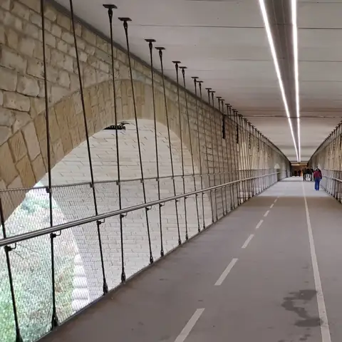 Langer, hell beleuchteter Steg mit markiertem Radweg zwischen den Steinbögen einer Brücke