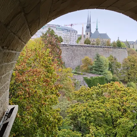 Blick durch einen Steinbogen auf bewaldete Hänge, Festungsmauern und die Türme einer Kathedrale