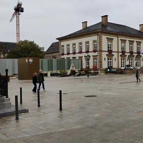 Weiter gepflasterter Platz vor einem hellen, lang gestreckten Gebäude mit Walmdach und Blumenkästen an den Fenstern