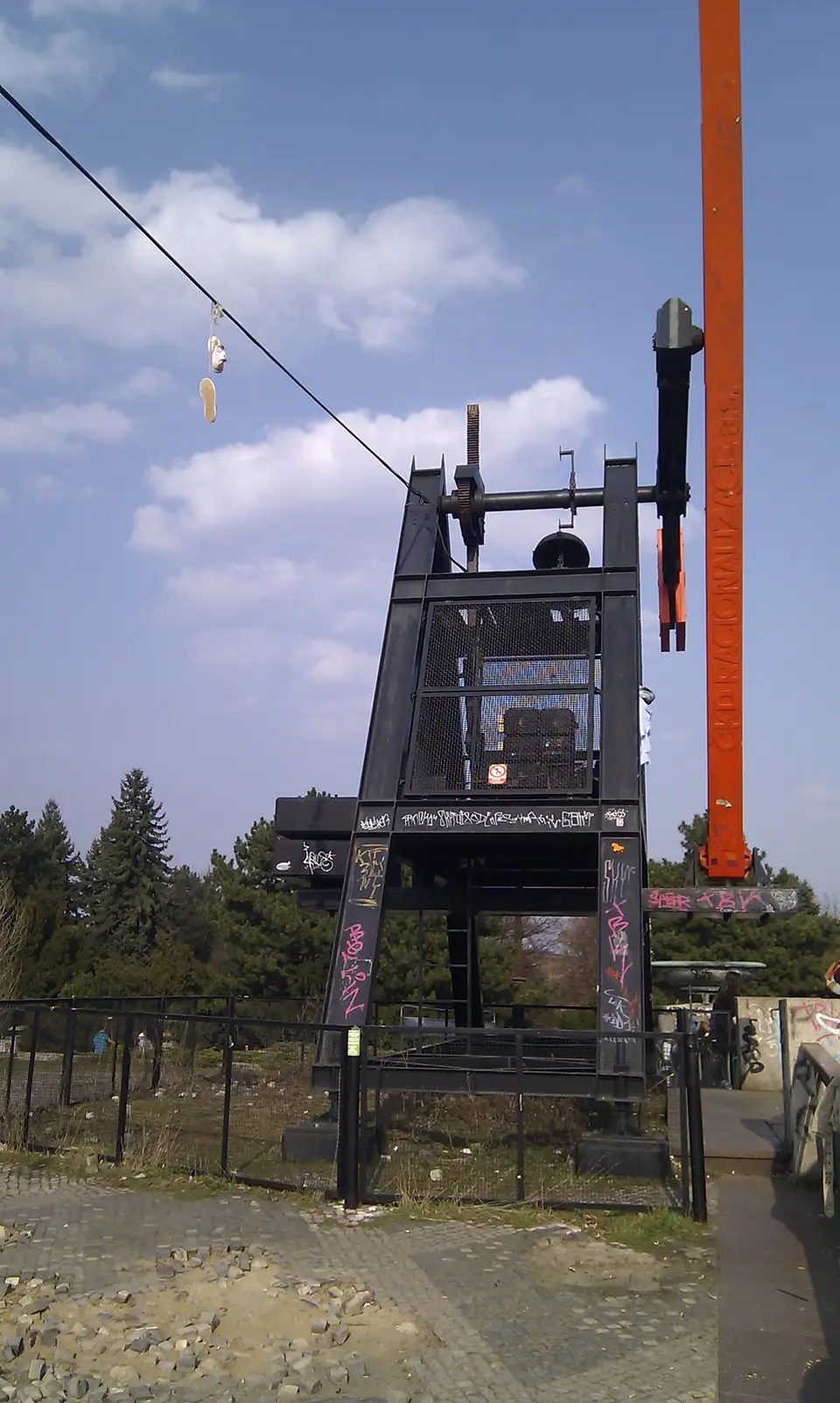 Dunkles Gestell des Prager Metronoms mit hohem orangefarbenem Pendelarm vor blauem Himmel