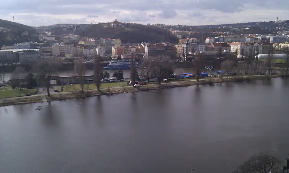 Blick von einer Anhöhe über die Moldau auf eine baumbestandene Uferzone und die Häuser von Smíchov