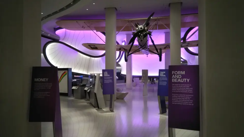 Mathematikgalerie im Science Museum mit großen geschwungenen, violett beleuchteten Deckenelementen, Vitrinen und einem aufgehängten Flugzeug