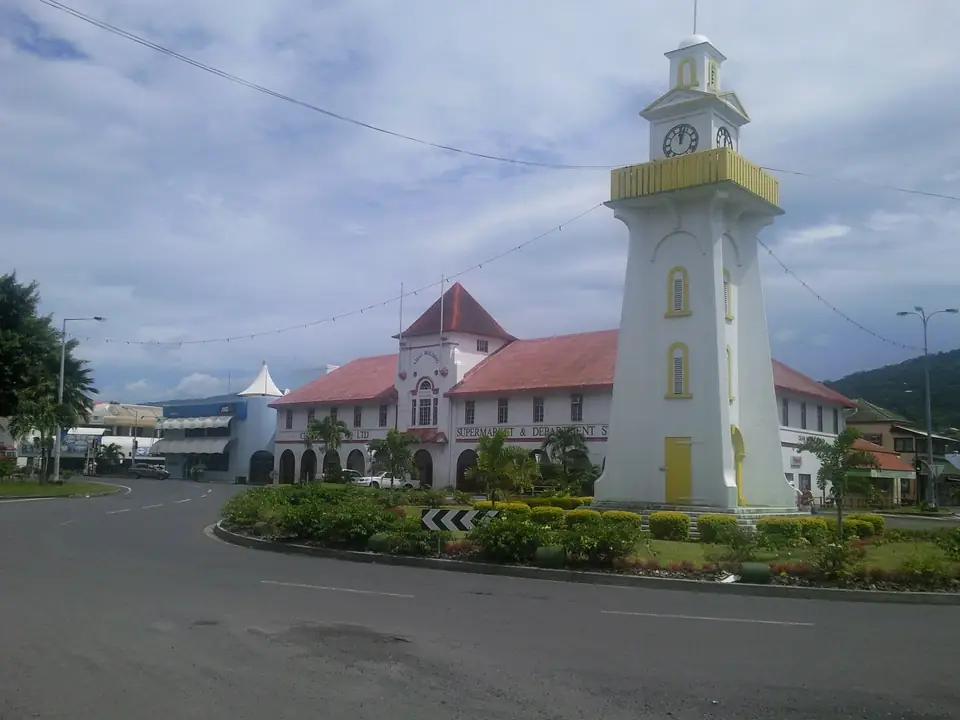 Weißer Uhrturm mit gelben Verzierungen auf einer Verkehrsinsel in Apia, dahinter Gebäude mit roten Dächern