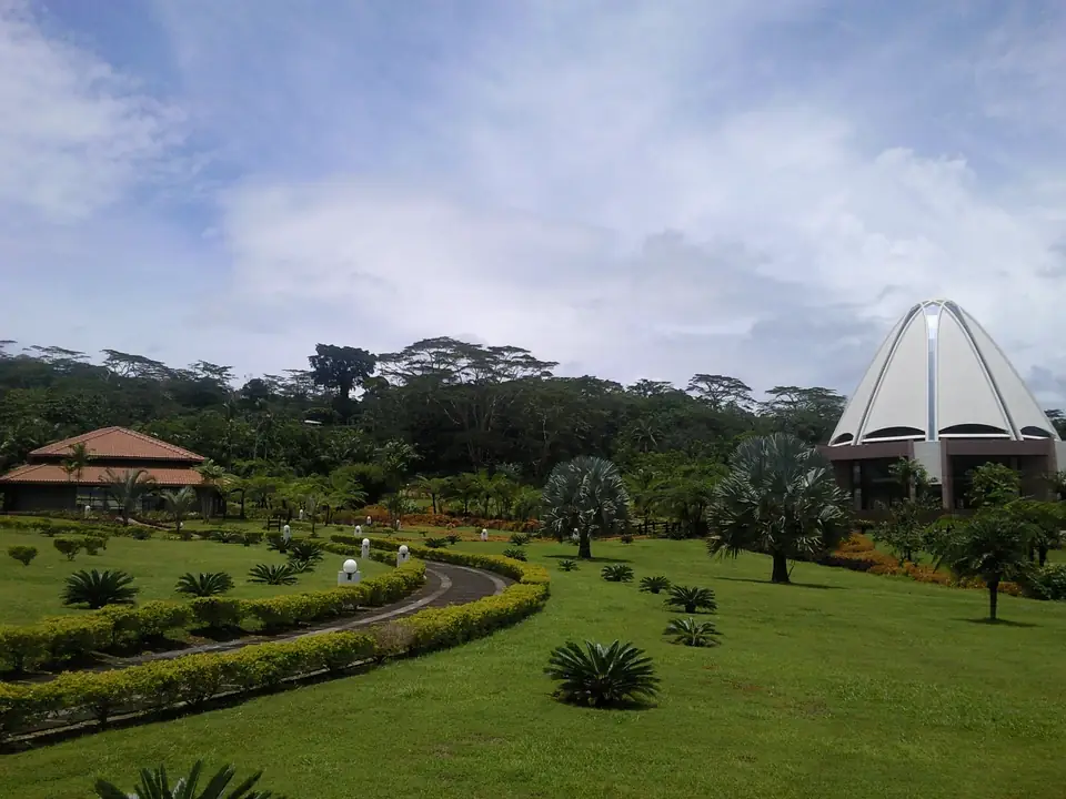 Heller Bahá’í-Tempel mit spitz zulaufender Kuppel hinter geschwungenen Gartenwegen und niedrigen Hecken