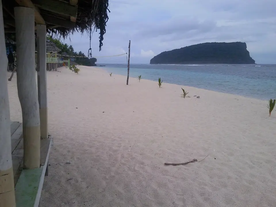Heller Strand von Lalomanu mit offenen Fales, Volleyballnetz, jungen Palmen und einer grünen Insel vor der Küste